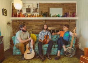 The three members of Tall Poppy String Band sit on a couch holding their instruments
