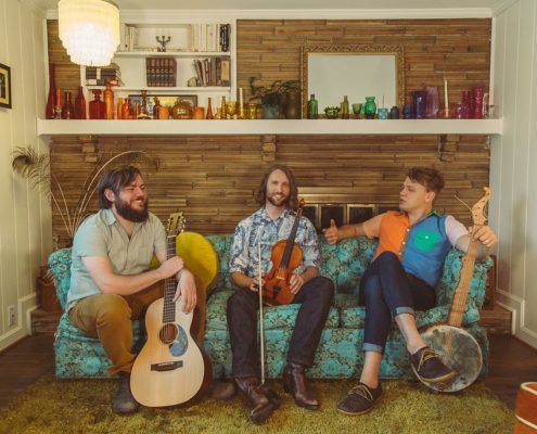 The three members of Tall Poppy String Band sit on a couch holding their instruments