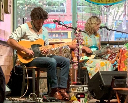 Two musicians sit on small stage holding guitars