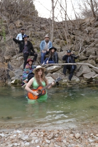 woman with a guitar half-immersed in a river. people on the shore look on